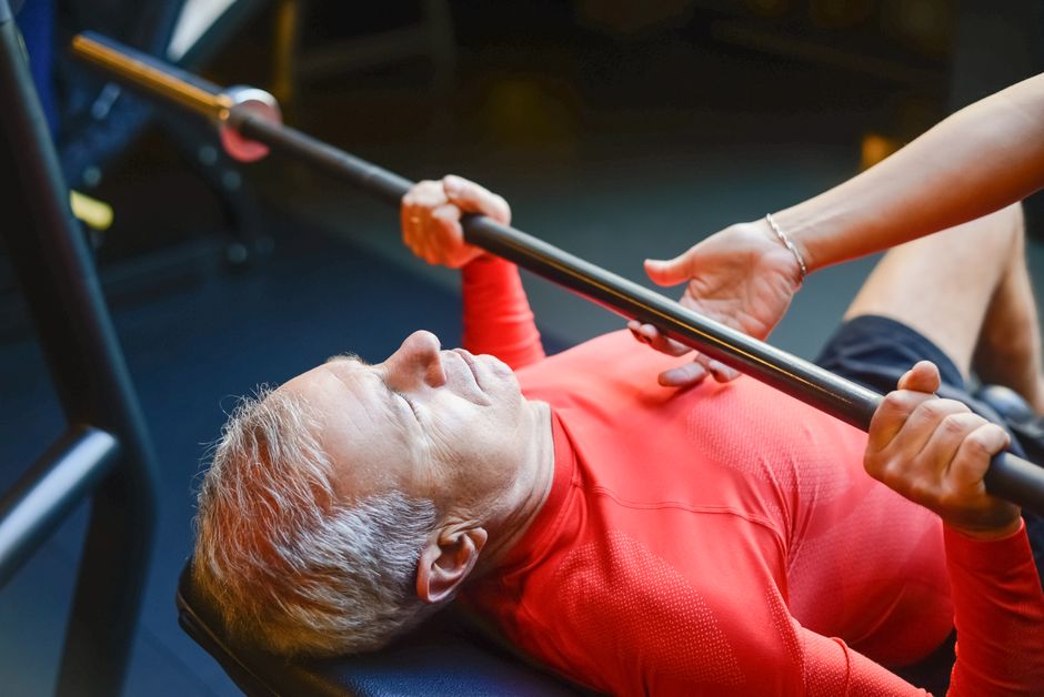 An elderly man performs a bench press exercise with a trainer in a gym